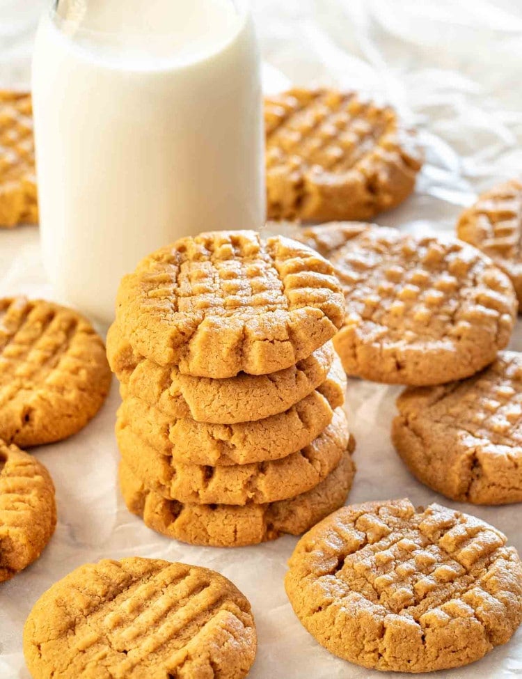 a stack of 5 peanut butter cookies with a glass of milk in the background surrounded by more cookies.