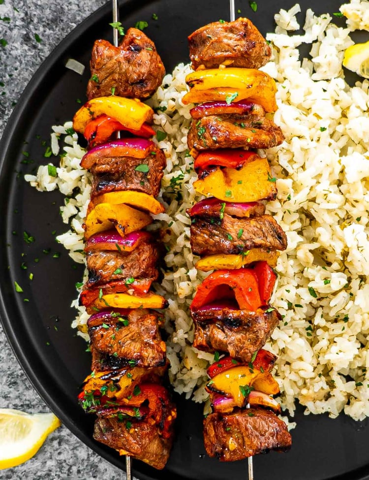 2 skewers of beef shish kebabs on a plate next to a bed of rice on a black plate.