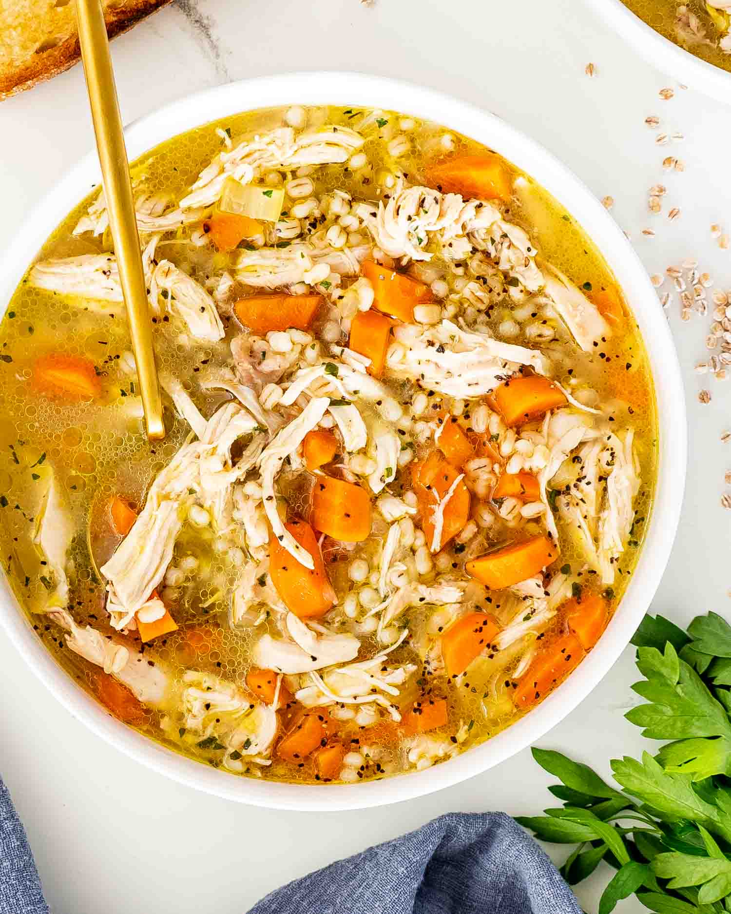 freshly made chicken barley soup in a white bowl with a golden spoon in it.