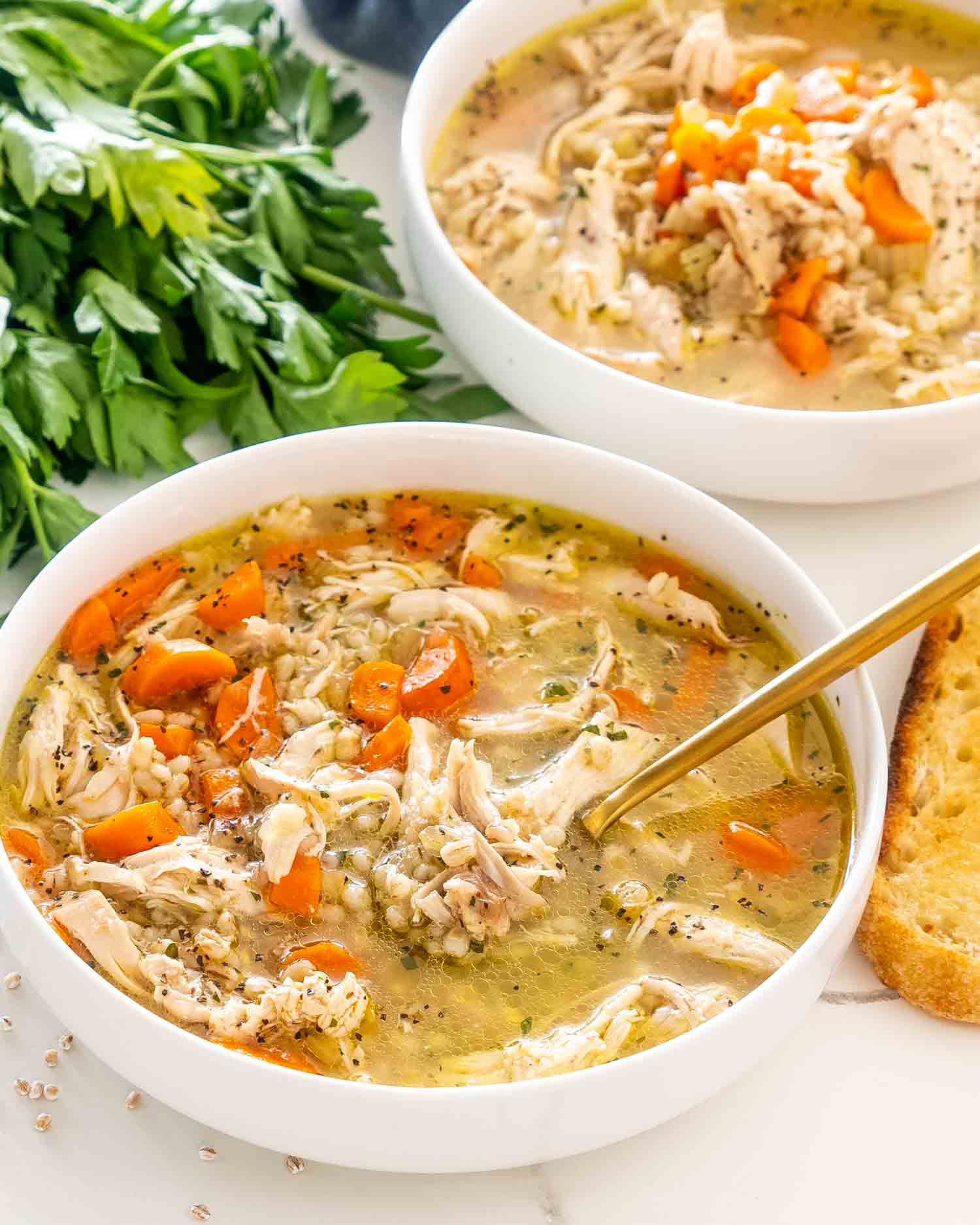 freshly made chicken barley soup in a white bowl with a golden spoon in it.