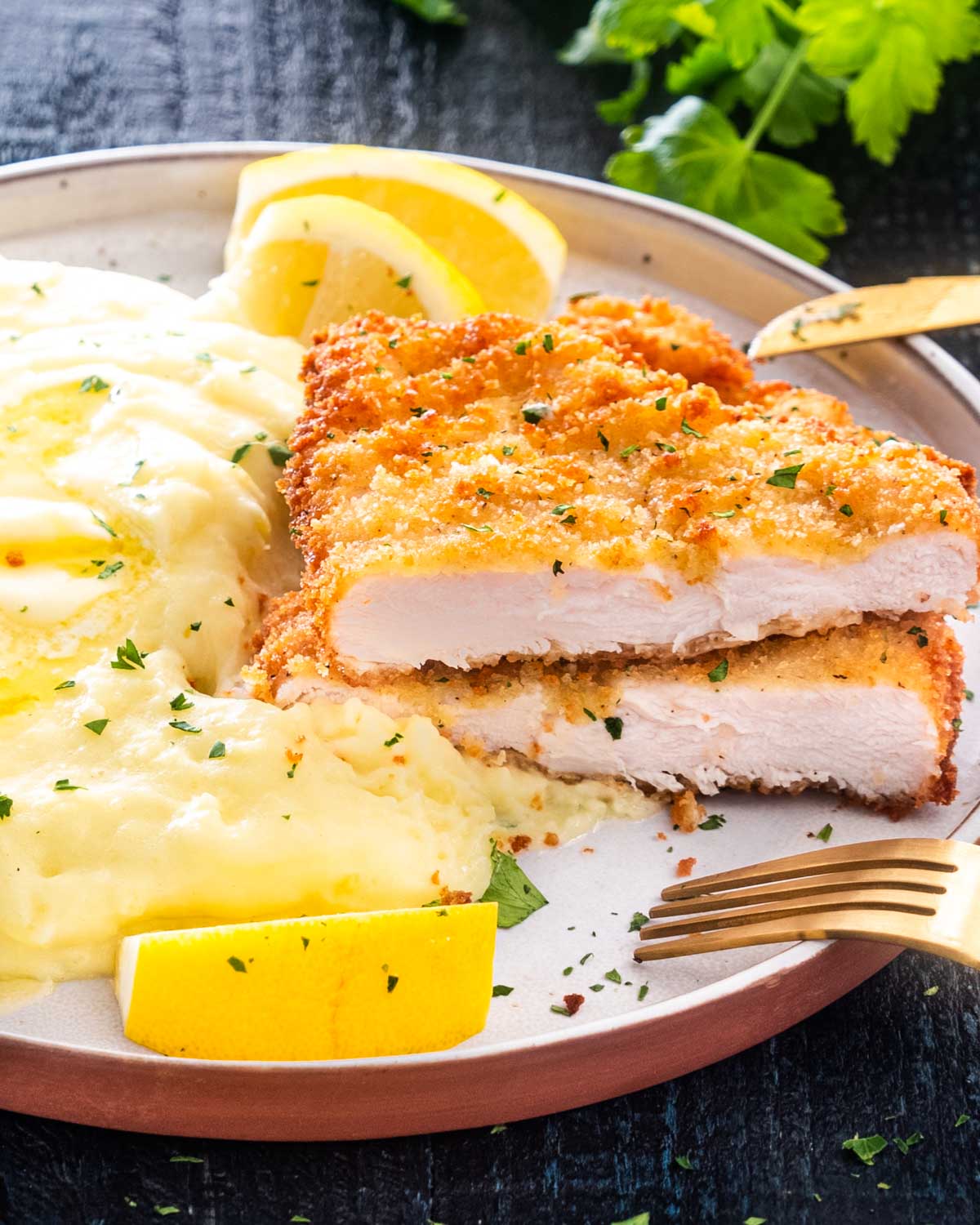 a chicken schnitzel on a plate with mashed potatoes and lemon wedges.
