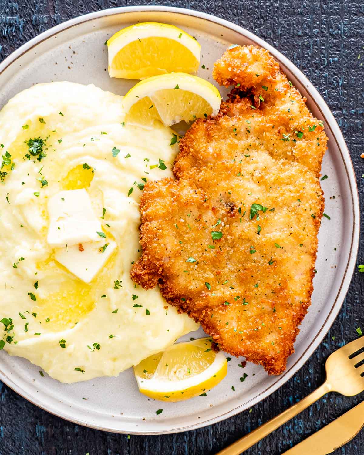 a chicken schnitzel on a plate with mashed potatoes and lemon wedges.