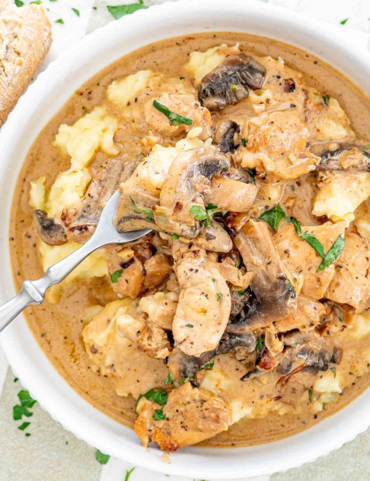 A bowl of chicken stroganoff with mushrooms atop smooth mashed potatoes, sprinkled with herbs.