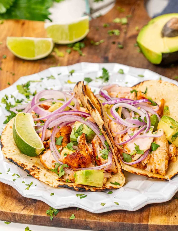 two chicken tacos with avocados and red onion on a white plate.