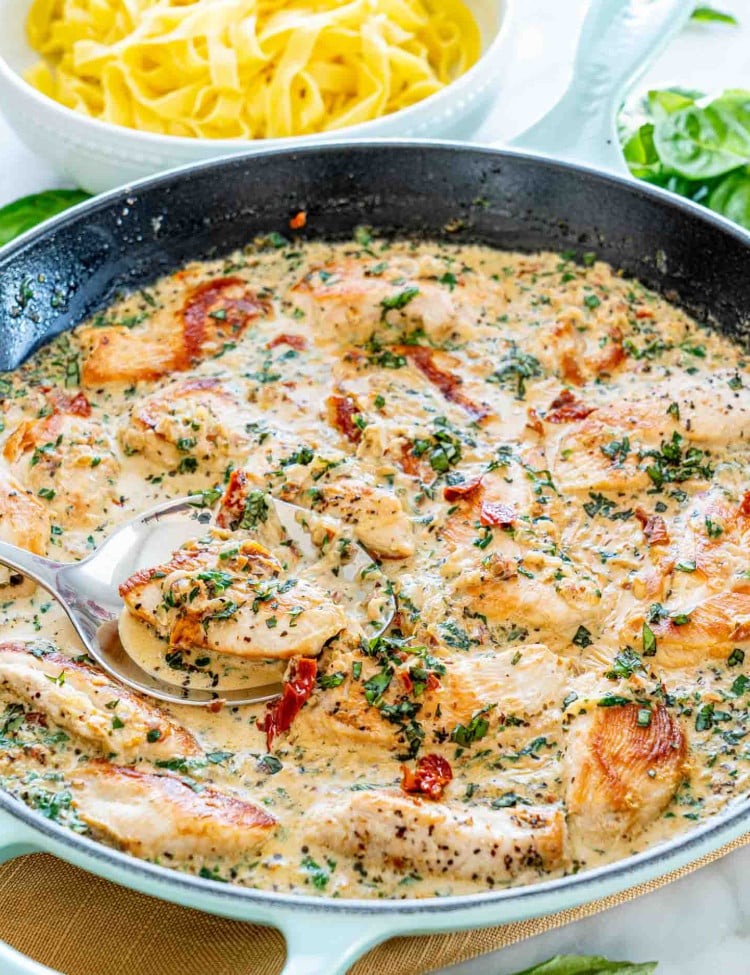 side view shot of creamy tuscan chicken in a blue skillet with a plate of noodles in the background.