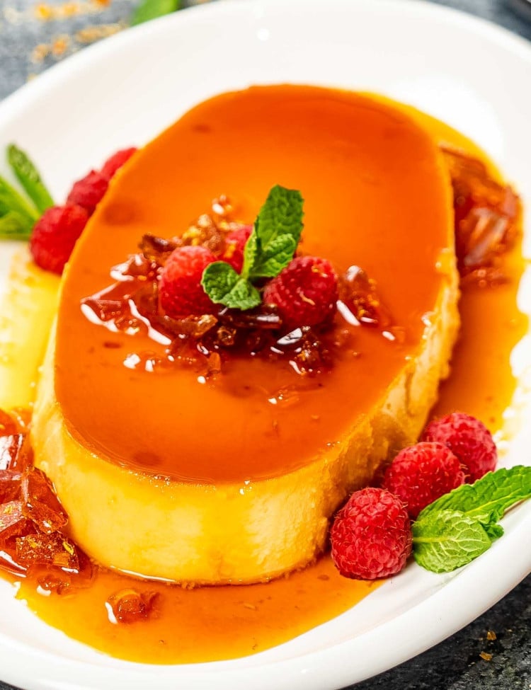 a gorgeous creamy flan on a white oval plate garnished with raspberries.