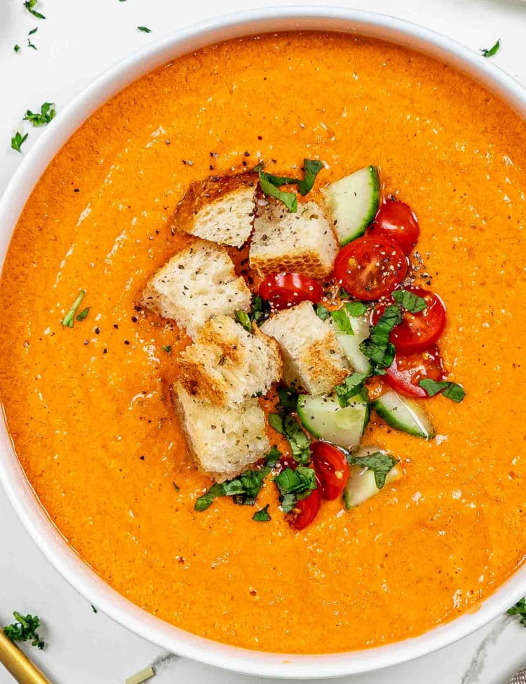 a white bowl loaded with fresh gazpacho and topped with croutons, chopped tomatoes and cucumber and a sprinkle of basil.