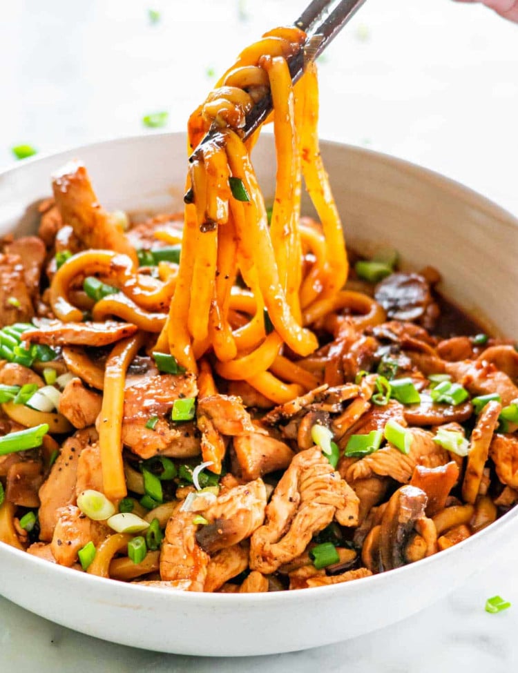 a hand pulling up some noodles with chop sticks from a plate full of hoisin chicken with udon noodles