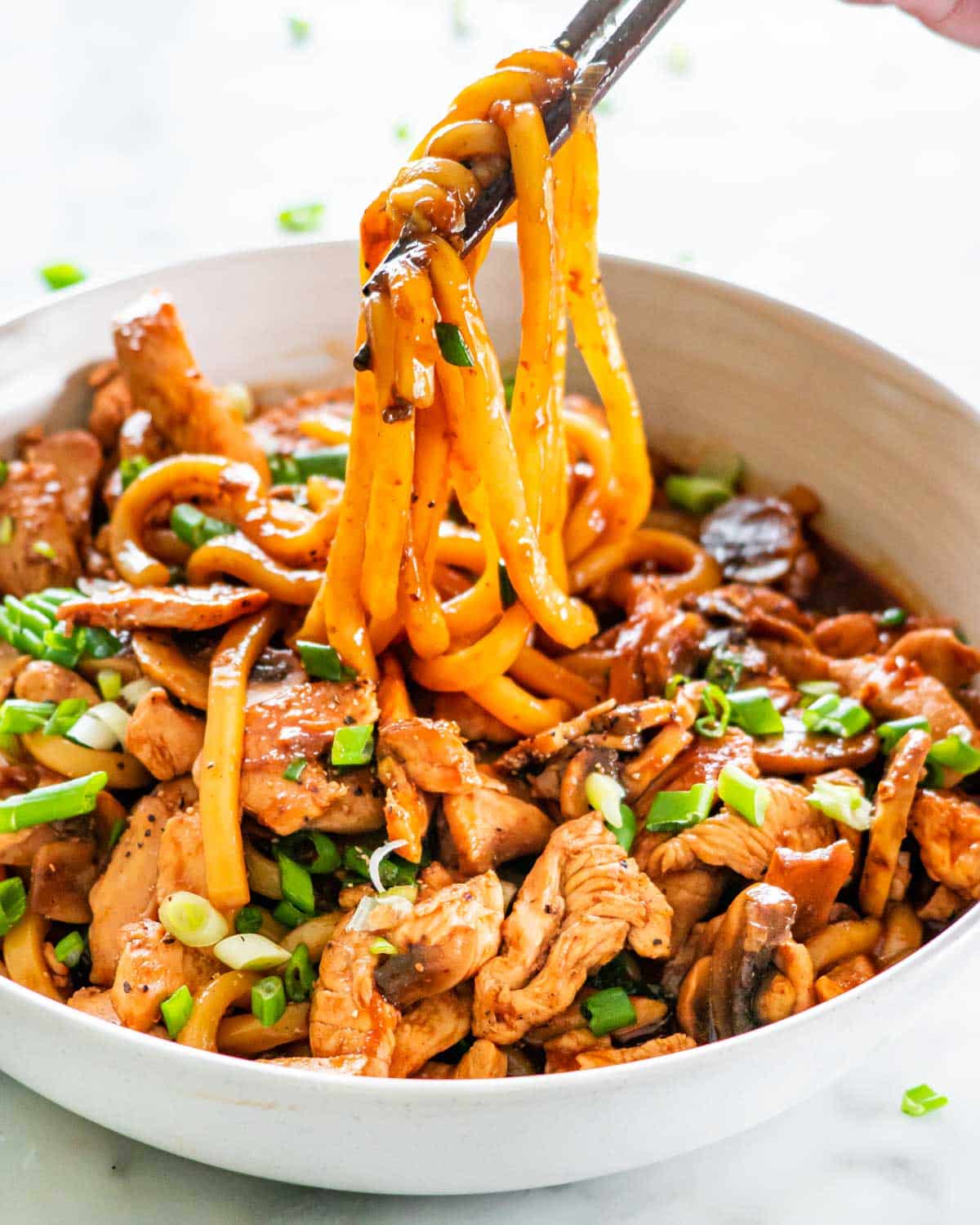 a hand pulling up some noodles with chop sticks from a plate full of hoisin chicken with udon noodles