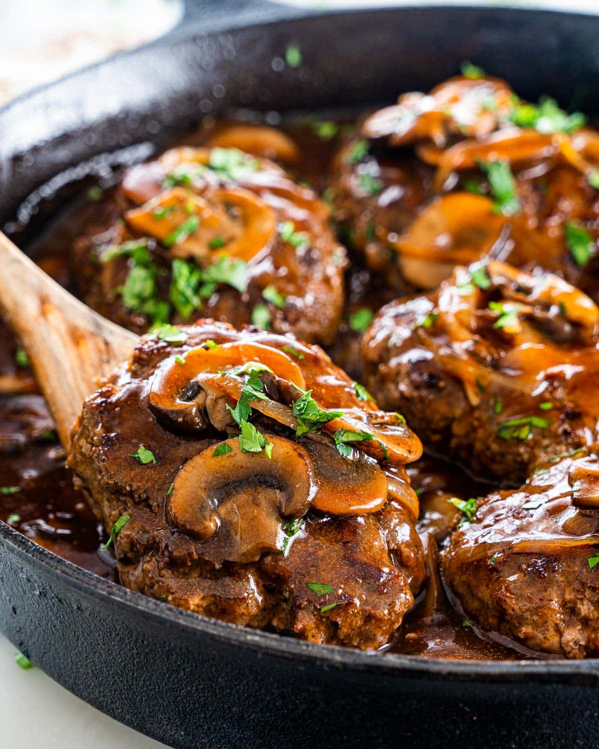 a serving spoon lifting a salisbury steak in a black skillet full of salisbury steak