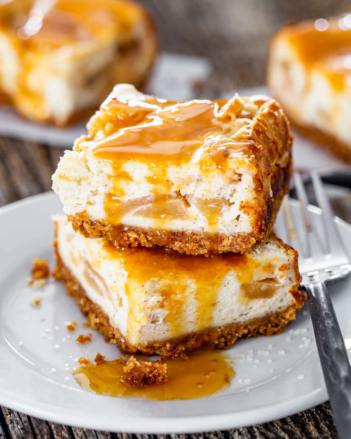 close up shot of two caramel apple pie cheesecake bars covered in caramel sauce stacked on top of one another on a plate